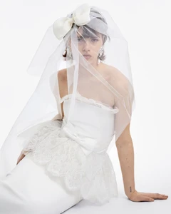 Model wearing a white strapless dress and tulle veil with a large bow against a plain white background