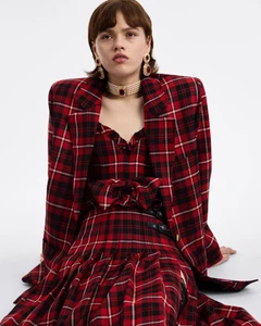 Model Ayla Murray seated in a red and black tartan plaid blazer and pleated dress with a pearl choker against a white studio backdrop.