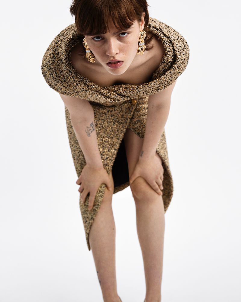 Model leaning forward wearing a textured tweed dress and ornate gold earrings against a white background