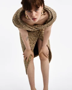 Model leaning forward wearing a textured tweed dress and ornate gold earrings against a white background