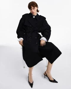 Model seated on a transparent chair wearing a black belted trench coat and black slingback heels against a white background