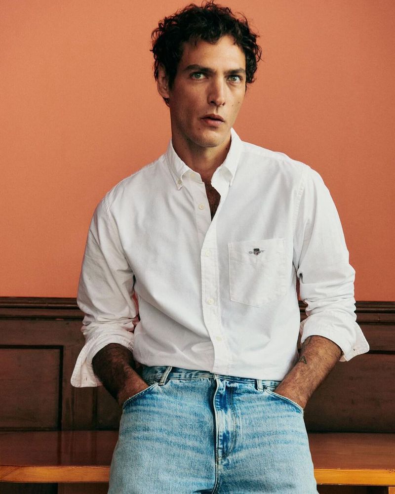 Studio shot of a dark-haired man in a white button-down shirt and blue jeans standing indoors against an orange-colored backdrop.