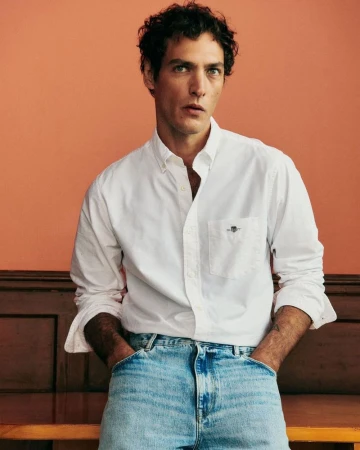 Studio shot of a dark-haired man in a white button-down shirt and blue jeans standing indoors against an orange-colored backdrop.