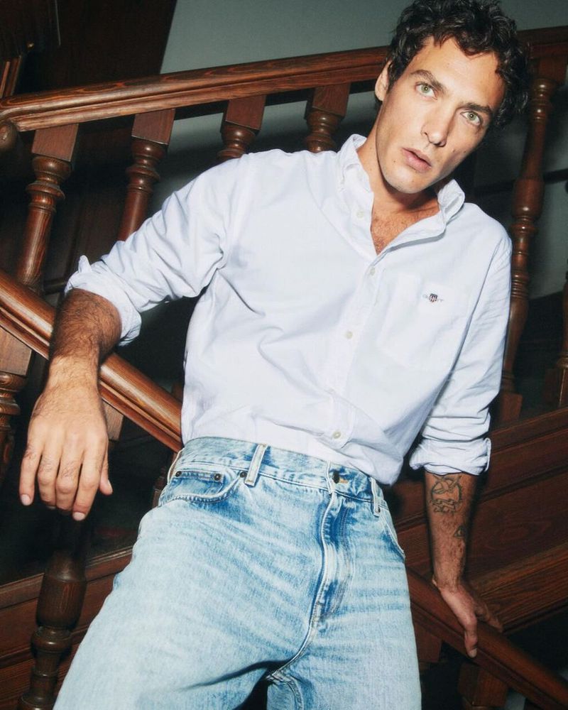 A young adult male is captured indoors, leaning on a wooden staircase, wearing a white collared shirt and light blue denim jeans.