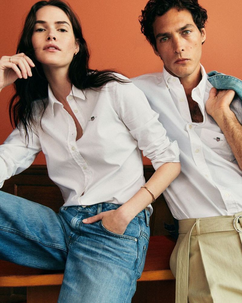 Two models in white shirts and jeans, seated indoors against an orange background, captured in a medium shot for a fashion editorial.
