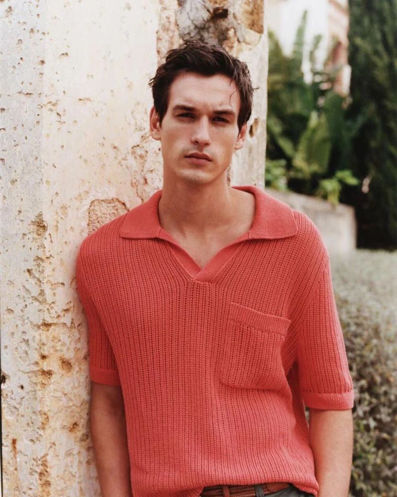 Portrait of a male model in a coral, ribbed knit polo shirt leaning against a textured beige wall with greenery visible in the background.