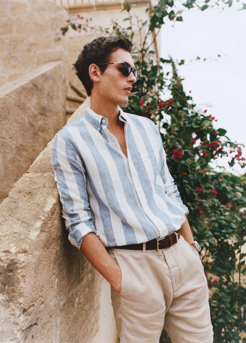 Man in striped shirt leans against wall outdoors. He wears sunglasses, and plants with red flowers are behind him in soft daylight.
