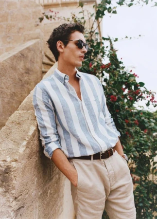 Man in striped shirt leans against wall outdoors. He wears sunglasses, and plants with red flowers are behind him in soft daylight.