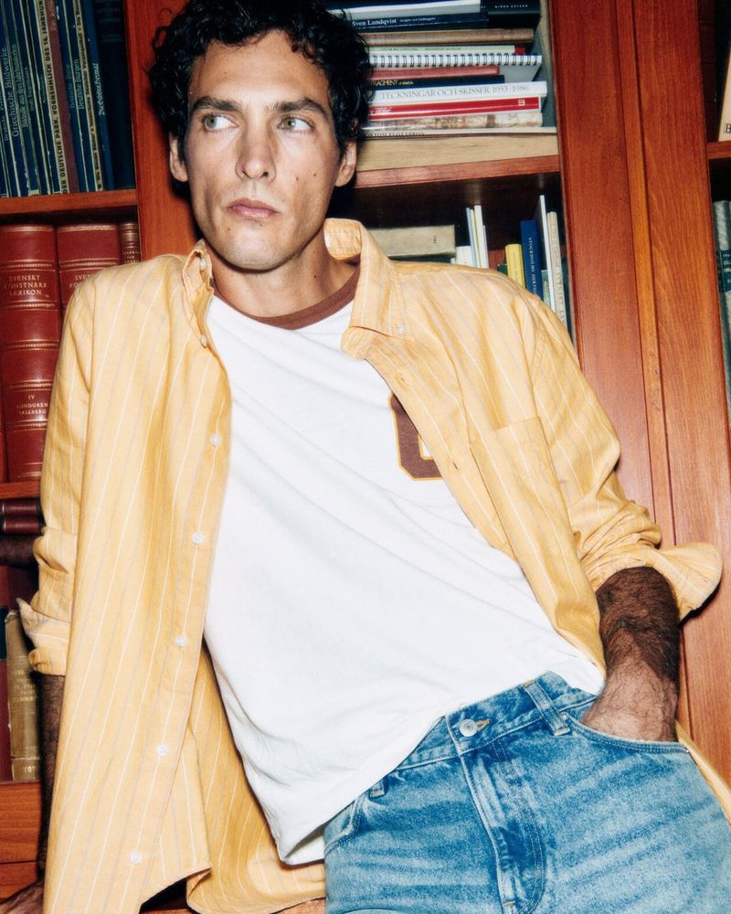 Attractive man in a white t-shirt and yellow shirt, leaning casually against a bookshelf in a cozy, indoor setting, medium shot.