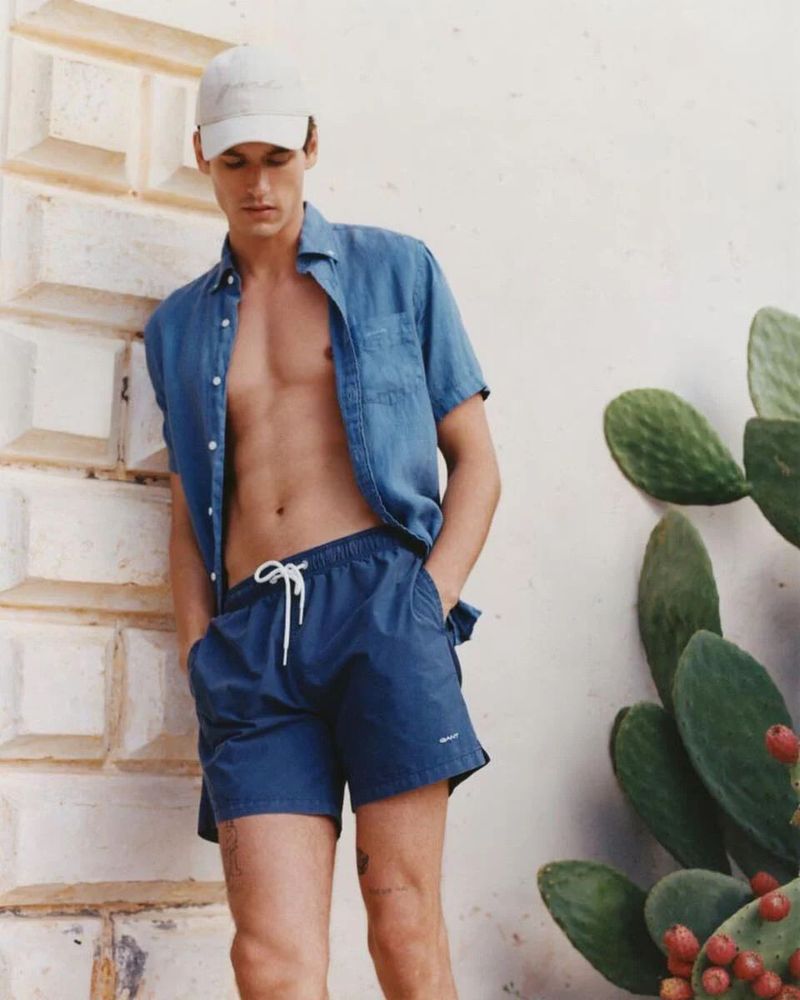 Male model posing outdoors with open denim shirt and blue shorts, leaning against wall with cactus plant in the background.