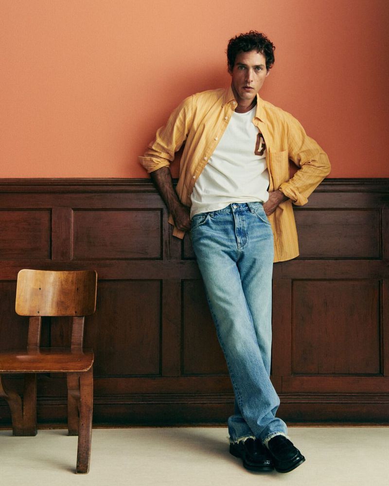 Fashionable adult male poses in front of a paneled wall, wearing a casual shirt, t-shirt, and jeans, with a vintage wooden chair nearby.