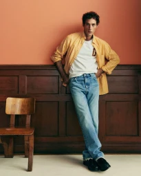 Man wearing yellow shirt and blue jeans leaning against a wooden panelled wall beside a chair