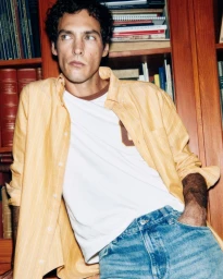 Male model in a yellow striped shirt over a white t-shirt and blue jeans standing in front of a wooden bookcase.