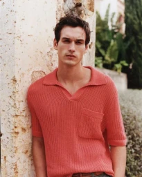 Male model wearing a coral red knit polo shirt leaning against an exterior textured stone wall for Gant