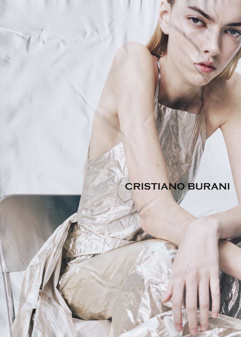 A fair-skinned fashion model poses while sitting on a metal chair in a shiny silver halter dress with dramatic studio lighting and a silver backdrop.