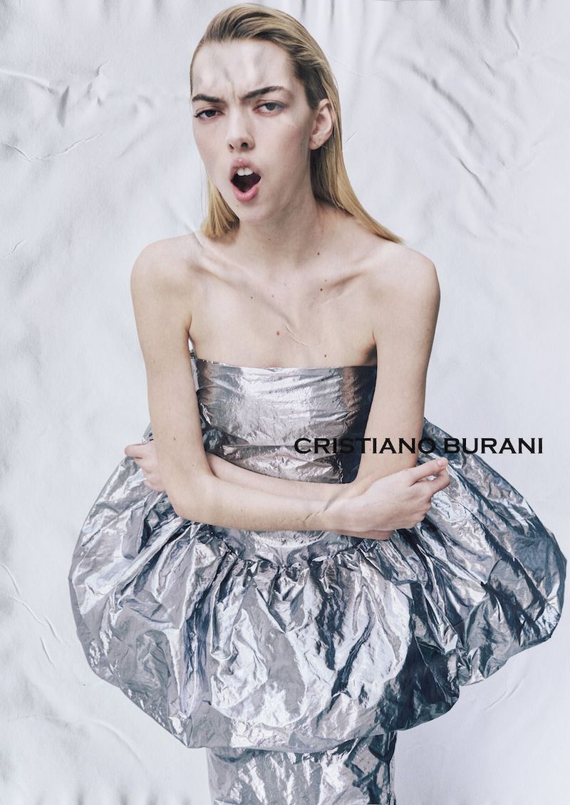 A blonde female fashion model with mouth open wears a silver foil dress against a textured white background in a studio shot.