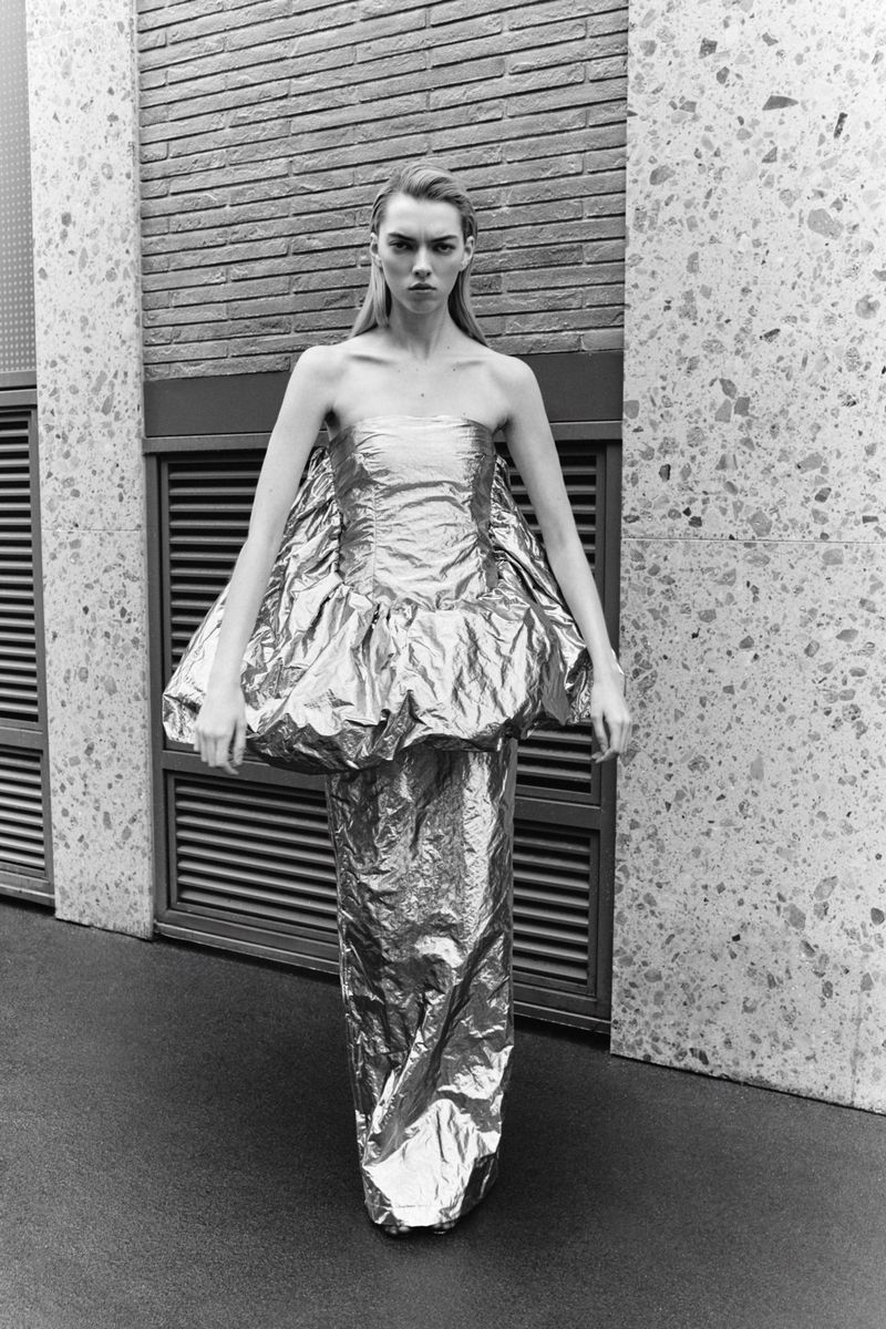 Monochrome portrait of a fashion model in a silver metallic dress standing in front of a gray brick and granite wall, eyes forward.