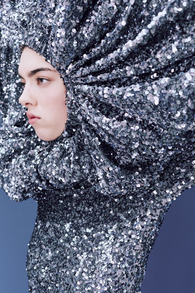 Adult fashion model in a silver sequin garment with a hood, photographed in a studio setting against a dark blue background.