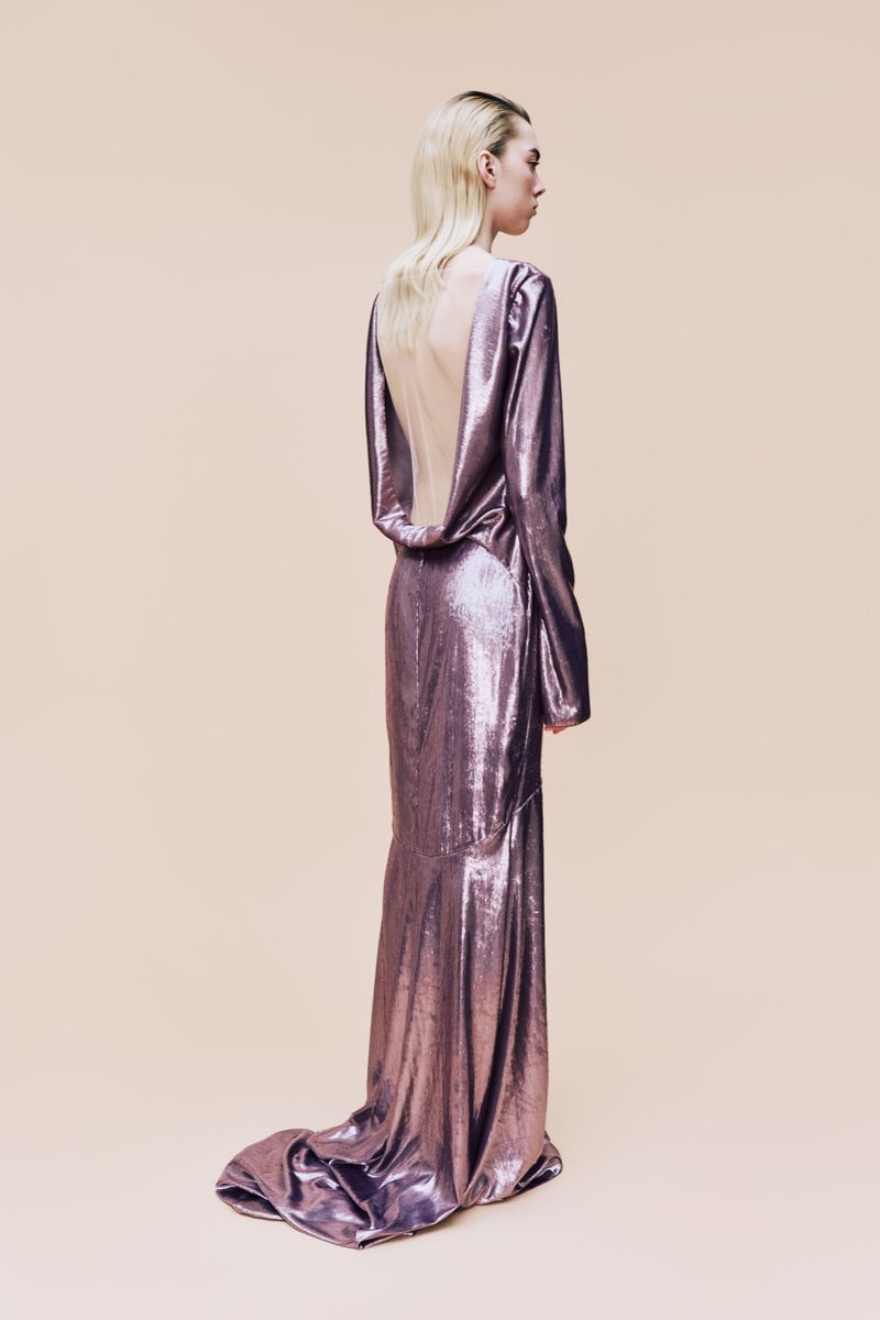 A blonde woman, seen from the rear, poses in a shiny, long-sleeved gown against a pale peach backdrop in a fashion or beauty context.