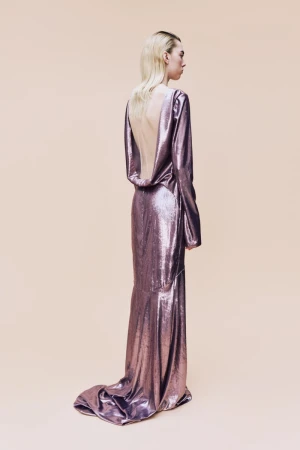 A blonde woman, seen from the rear, poses in a shiny, long-sleeved gown against a pale peach backdrop in a fashion or beauty context.