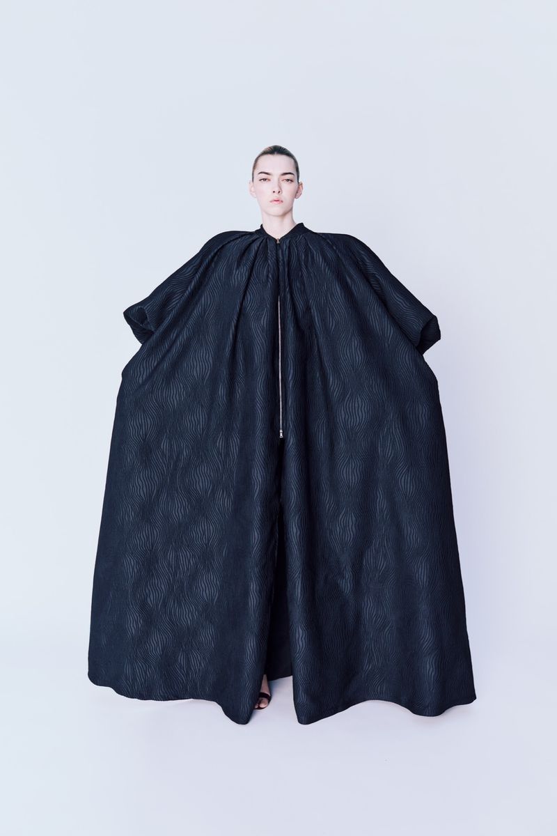 Woman stands wearing an oversized black coat with a zipper in a full shot against a bright white backdrop in a fashion photography style.