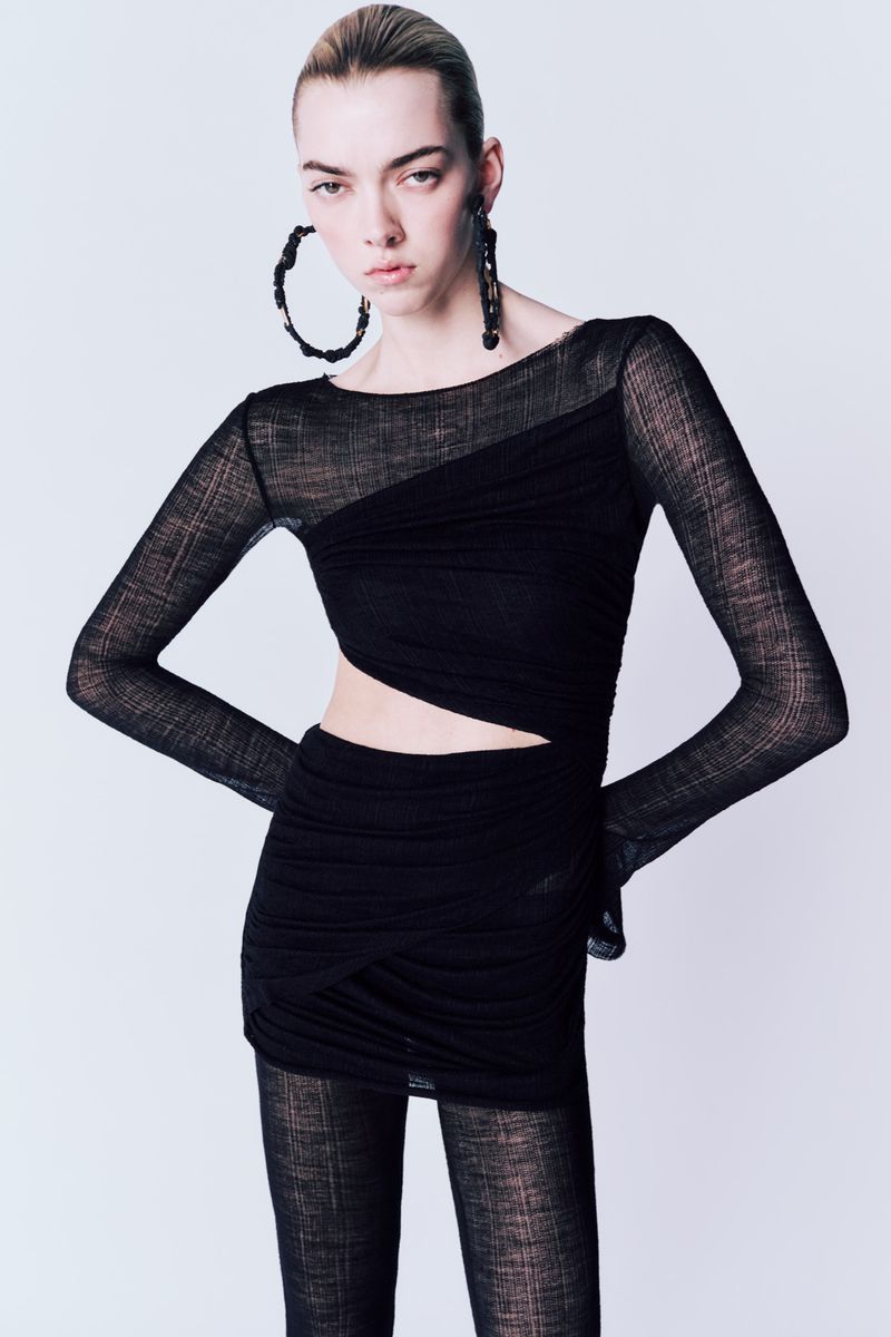 Fashion model poses in a black long-sleeved mini dress with geometric cutouts and oversized earrings, against a light grey backdrop.
