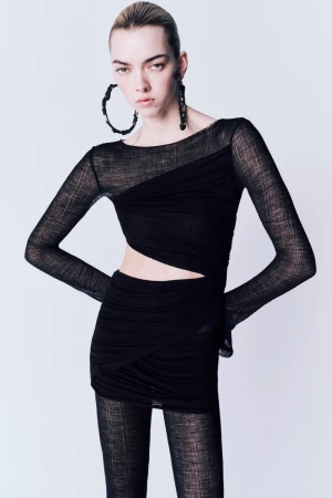 Fashion model poses in a black long-sleeved mini dress with geometric cutouts and oversized earrings, against a light grey backdrop.