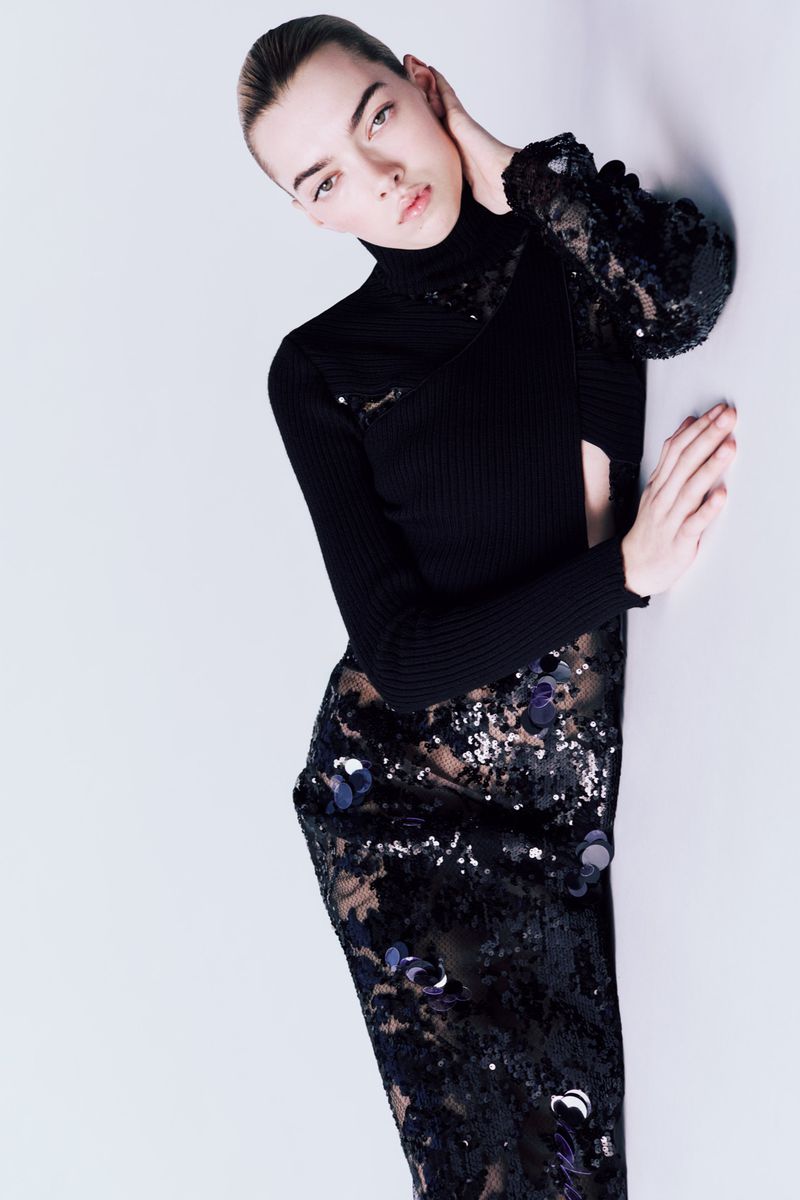 Fashionable woman in a black cut-out turtleneck and sequined skirt poses against a white wall in a studio shot.