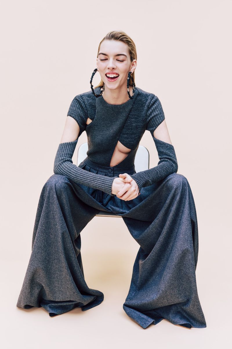 Blonde woman in a grey knit outfit with flared pants sitting on a chair with hands clasped against a beige background looking cheerful.