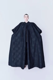 Model wearing an oversized voluminous black structured cape coat against a pale studio seamless backdrop.