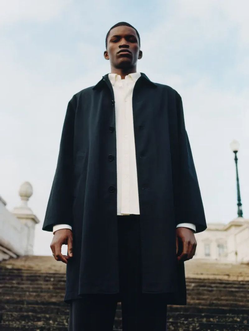 An African American male model poses in a dark coat and white shirt on a staircase under a cloudy sky, showcasing a fashion-forward editorial style.