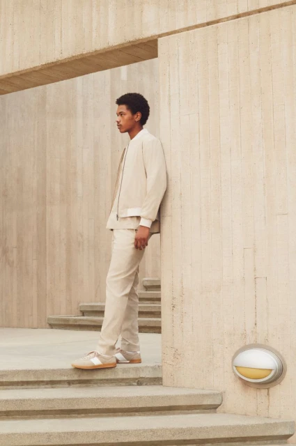 Young adult man in beige jacket and pants poses on steps of a concrete building with a neutral expression on his face in a medium shot.