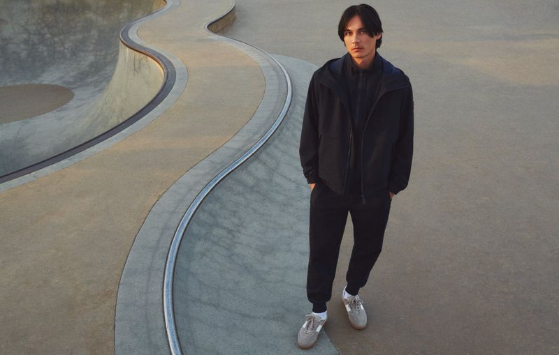 Adult male in jacket and sweatpants stands at edge of concrete skatepark bowl in daylight. Concrete bowl curves behind him.