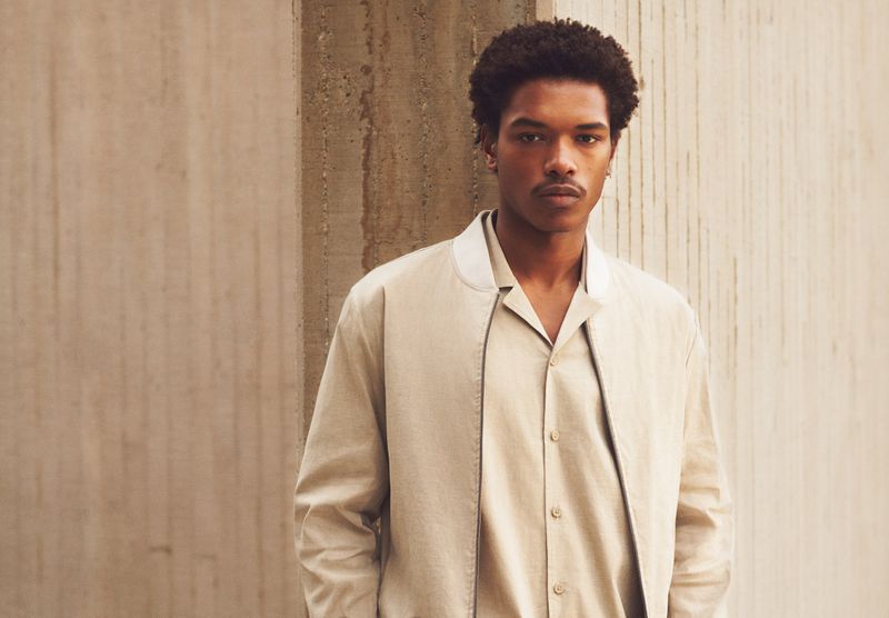 Portrait of a young man in a beige jacket and matching shirt standing in front of a concrete wall, looking directly at the camera.