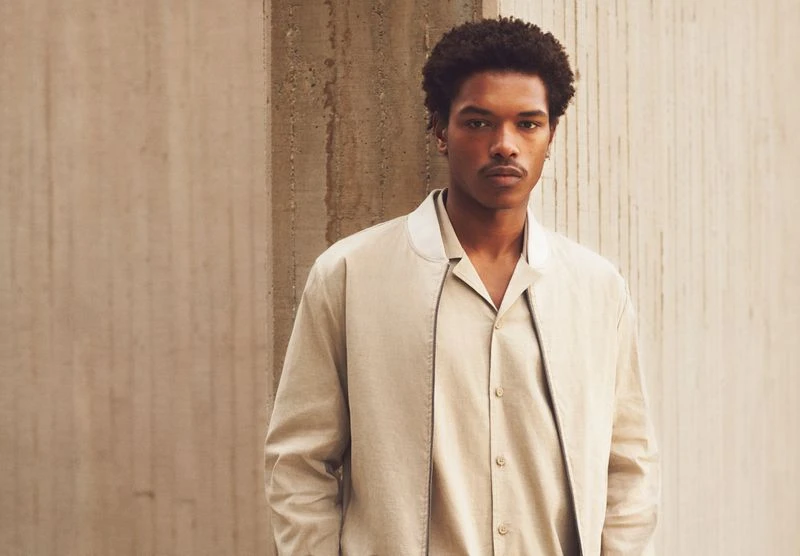 Portrait of a young man in a beige jacket and matching shirt standing in front of a concrete wall, looking directly at the camera.