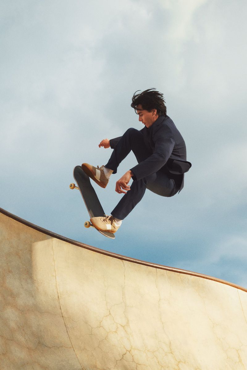 A skateboarder wearing a dark suit jumps off a concrete ramp in a skatepark against a partly cloudy sky during daylight hours.