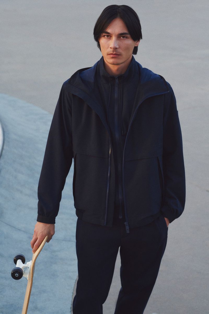Male model in navy hooded jacket and trousers holds a skateboard in a skatepark. Fashion shoot with neutral tones.