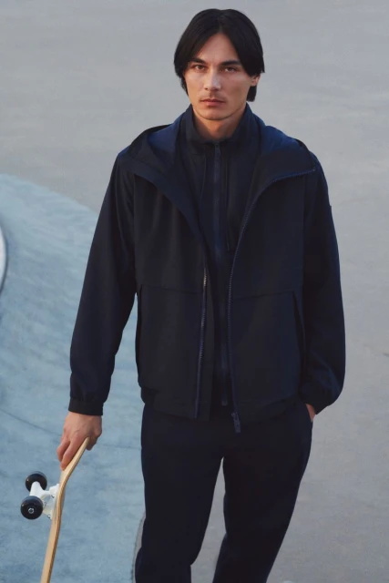 Male model in navy hooded jacket and trousers holds a skateboard in a skatepark. Fashion shoot with neutral tones.