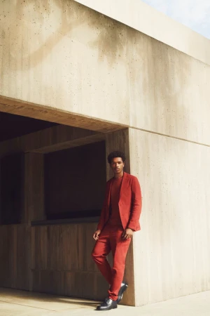 Man in a rust-colored suit leans against a tan concrete wall outdoors. Full-length shot, professional styling, fashion, modern architecture.