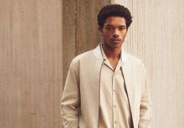 Young Black man with a mustache wearing a light beige button-down shirt and jacket against a concrete wall