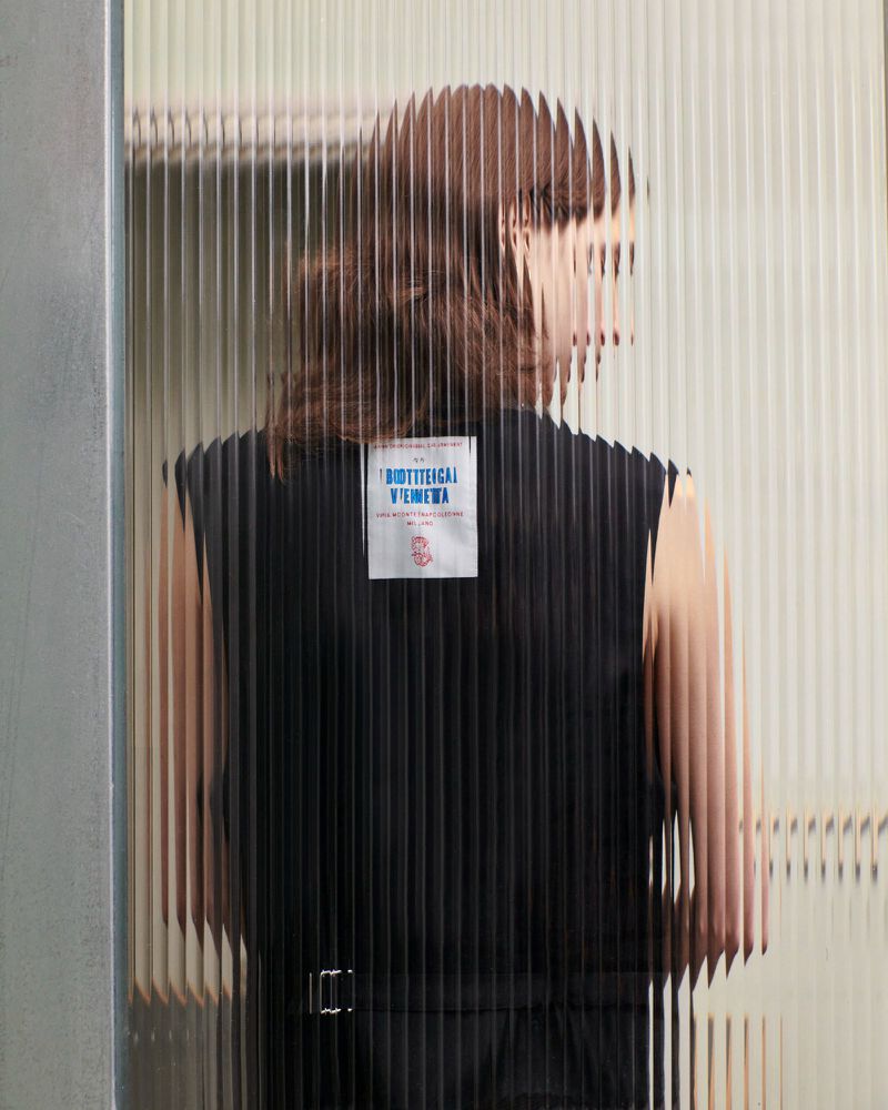 A person in a black sleeveless top is partially visible through a textured glass wall. The focus is on fashion and design.