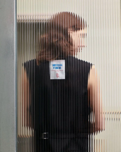 A person in a black sleeveless top is partially visible through a textured glass wall. The focus is on fashion and design.