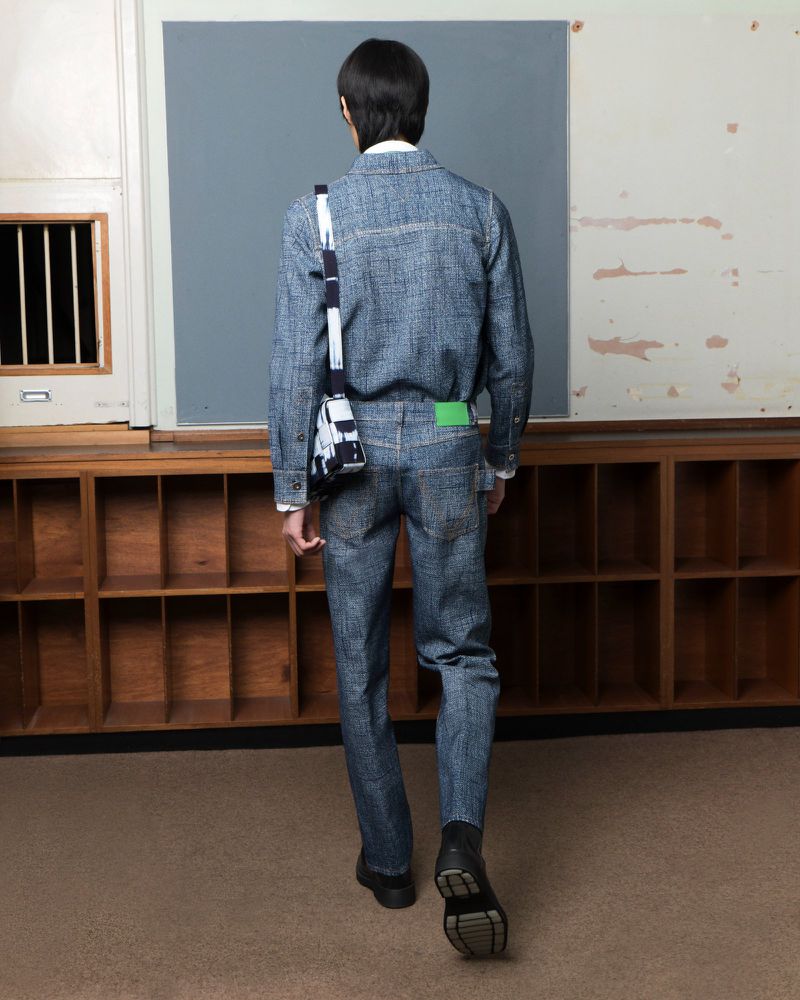 Rear view of a person walking in a classroom wearing a denim outfit and carrying a shoulder bag, near a window and wall.