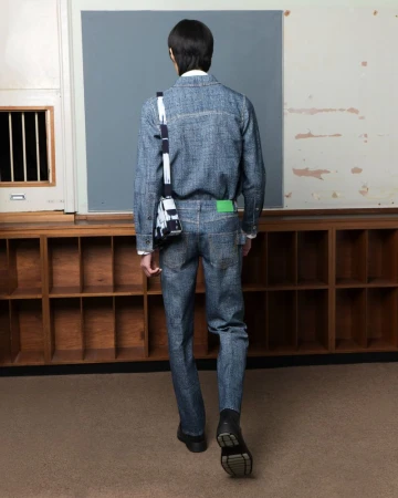 Rear view of a person walking in a classroom wearing a denim outfit and carrying a shoulder bag, near a window and wall.