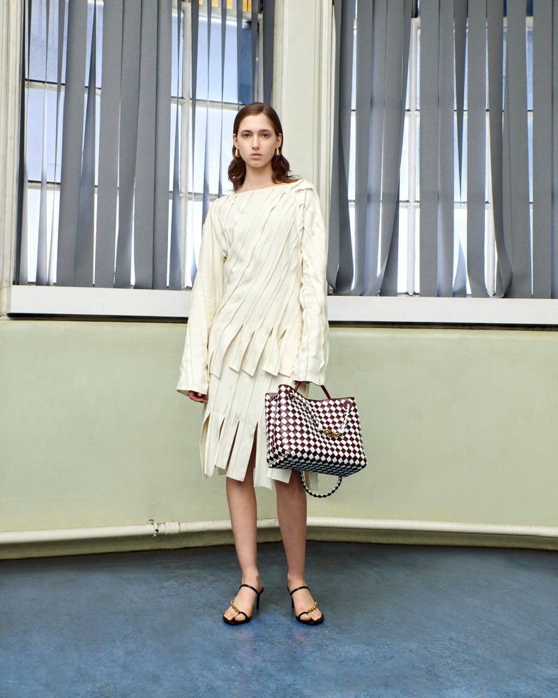 A woman in a cream-colored dress with a black and white tote bag stands in an indoor setting with natural lighting from a window.