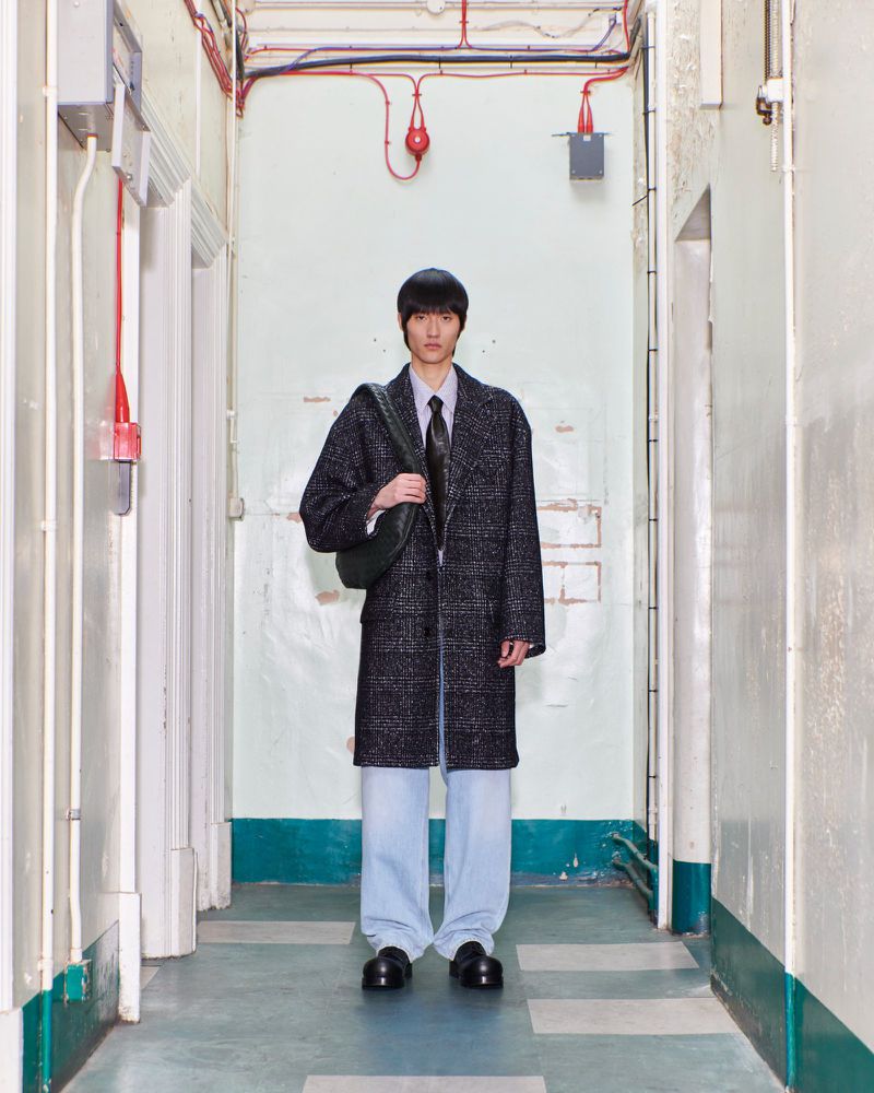 Young man in a coat stands in a hallway with a shoulder bag, wearing light blue trousers and dark shoes, lit by overhead lighting.