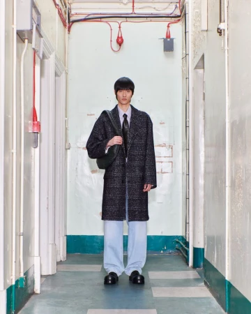 Young man in a coat stands in a hallway with a shoulder bag, wearing light blue trousers and dark shoes, lit by overhead lighting.