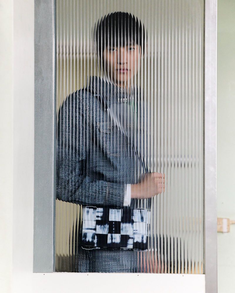 Portrait of an androgynous Asian person with short black hair, wearing a jacket and purse, standing behind a textured glass door.