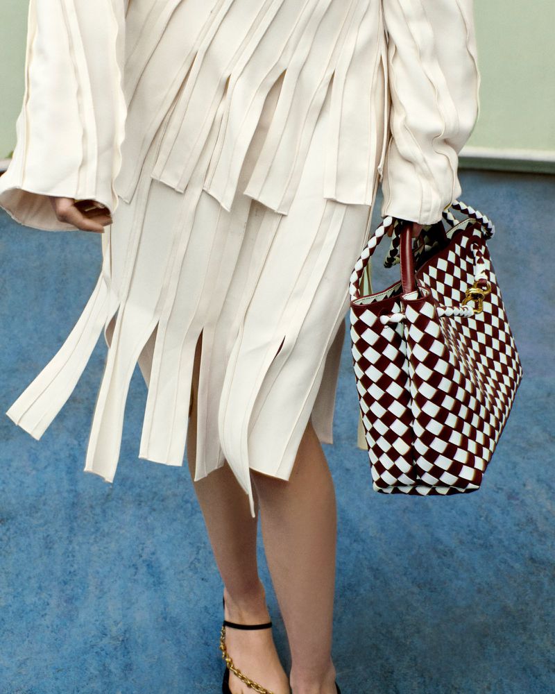 Cream-colored, asymmetrical skirt with fringe, held by a human, paired with strappy heels and a brown and white checkered purse.