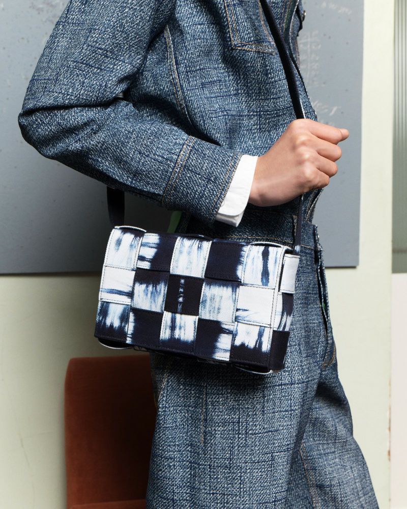 Fashionable woman showcasing a stylish, woven handbag with a black strap. She is wearing a denim jacket and pants.